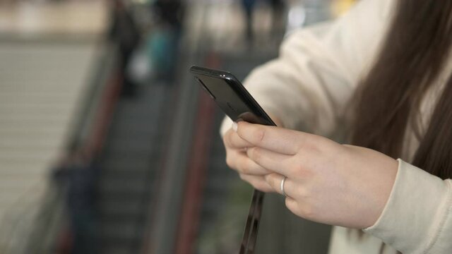 Hands of a young woman using a smartphone in a shopping mall. A young woman is holding a phone in a shopping mall in front of an escalator with people. Close up
