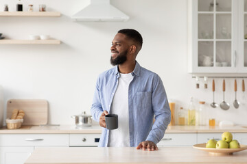 Smiling millennial black male with beard enjoys good morning and fresh coffee in cup and looks at free space
