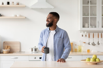Smiling millennial black male with beard enjoys good morning and fresh coffee in cup and looks at...