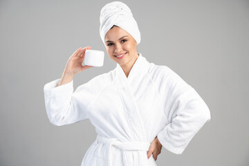 Portrait of young smiling caucasian woman in bathrobe holding jar with cosmetics cream near face and looking at camera. Beauty, skin care and advertising concept, horizontal shot