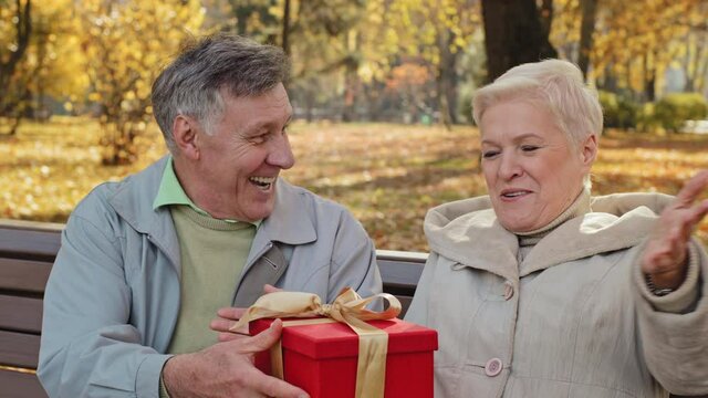 Mature man gives gift to beloved wife on birthday elderly woman happily laughs positive married couple celebrating anniversary unexpected surprise excited lady middle aged receive wrapped festive box
