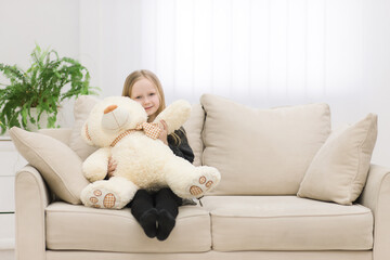 Photo of cute little girl sitting with teddy bear on white sofa.
