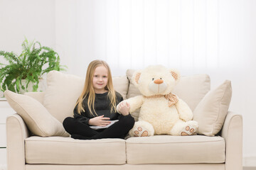 Photo of little girl and teddy bear together sitting on white sofa.
