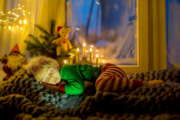 Cute toddler child in pajama, sitting on a fury blanket next to a window, looking outside at fireworks