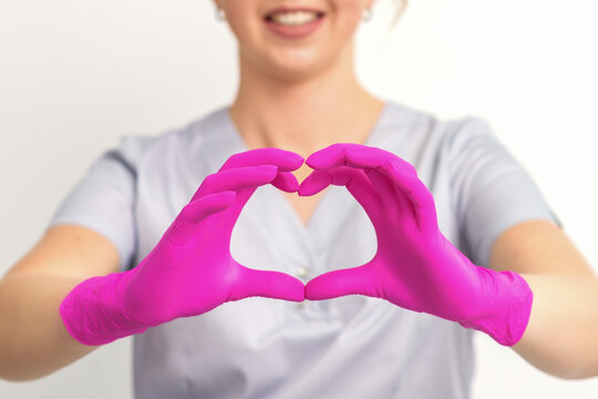 A smiling caucasian woman doctor wearing pink gloves in uniform showing the symbol of a heart against a white background