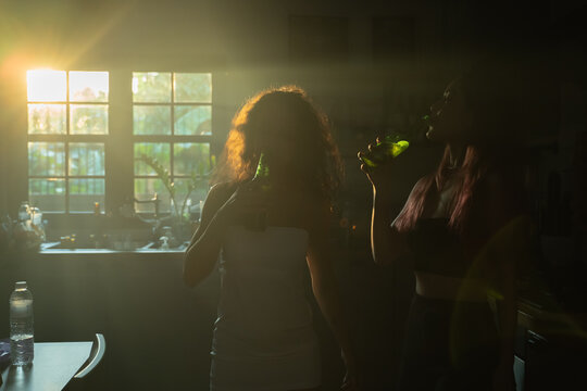 Group of two young woman friends drinking beer from bottle after late night party at home during sunrise while standing in kitchen