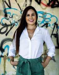 Young woman wearing green pants and a white shirt smiling in front of a graffiti wall of a railway station waiting for the next train. Mention of business travel and vacations.