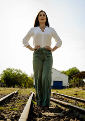 Young woman wearing green pants and a white shirt with serious expression and arms on her hips on a railway line waiting for the next train. Mention of business travel and vacations.