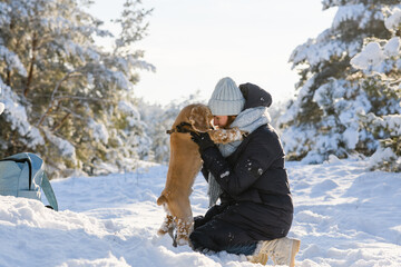 The girl sits on her knees in a snowdrift and the dog stands on its two hind legs. The girl hugs her pet. Young woman with American Cocker Spaniel in the winter forest.