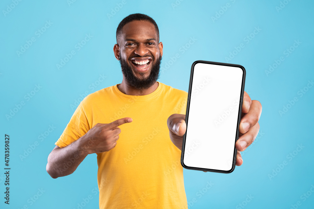 Poster handsome happy black guy pointing at smartphone with blank screen on blue background, mockup for web