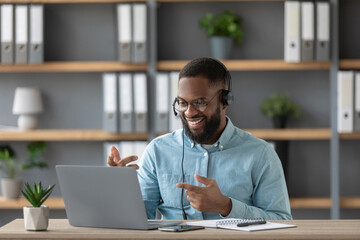 Smiling young black guy teacher or student with beard in glasses and headphones shows his fingers at pc screen