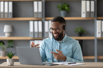 Obraz premium Smiling young black guy teacher or student with beard in glasses and headphones shows his fingers at pc screen