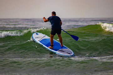 Surfer reitet mit eine Stand uo paddle (surfbrett) auf dem Meer über eine grße welle und könnte ins wasser fallen, er hält jedoch sein Gleichgewicht.   