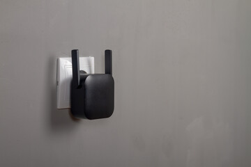 A black Wi-Fi signal amplifier is plugged into an outlet on a gray wall background. Copy Space