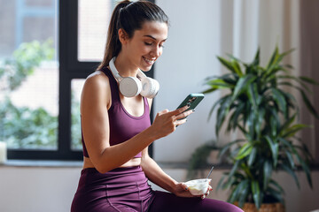 Sporty young woman eating a yogurt while using smartphone sitting on fitness ball at home.