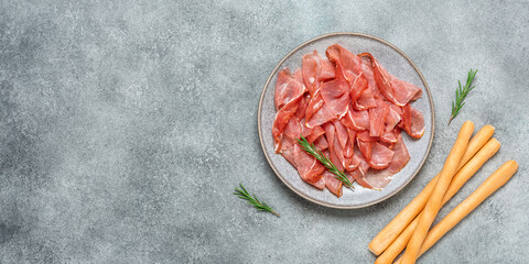 Slices of prosciutto di parma or jamon serrano in a plate and breadsticks on gray grunge background. Top view, flat lay, banner. - obrazy, fototapety, plakaty