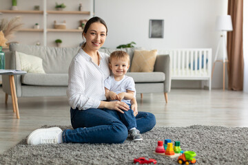 Happy caucasian mother and toddler son smiling at camera, playing together, sitting on floor carpet...
