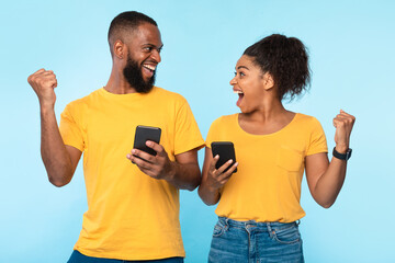 Online win, shopping sale concept. Excited millennial black couple using mobile phones, gesturing YES on blue background