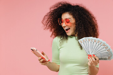 Young curly latin woman 20s years old wears mint t-shirt sunglasses using mobile cell phone hold...