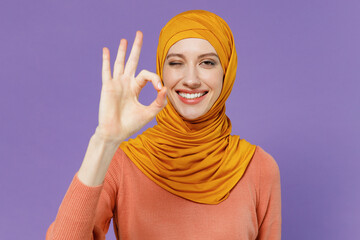 Smiling happy cheery young arabian asian muslim woman in abaya hijab yellow clothes showing okay ok zero finger gesture wink blink eye isolated on plain pastel light violet background studio portrait.