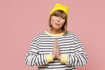 Elderly stylish woman 50s in striped shirt yellow hat hands folded in prayer gesture, begging about something isolated on plain pastel light pink background studio portrait. People lifestyle concept