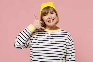 Elderly smiling stylish woman 50s wearing striped shirt yellow hat doing phone gesture like says call me back isolated on plain pastel light pink background studio portrait. People lifestyle concept.