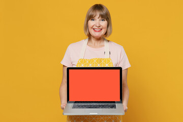 Elderly freelancer housekeeper housewife woman 50s in orange apron hold use work on laptop pc computer with blank screen workspace area isolated plain on yellow background. People household concept.