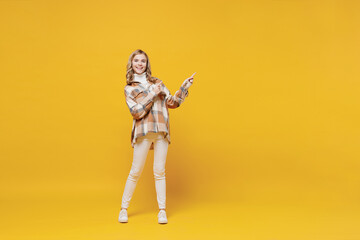Fototapeta na wymiar Full body little blonde smiling kid girl 13-14 years wearing checkered shirt point index finger aside on copy space area isolated on plain yellow background studio portrait. People lifestyle concept.