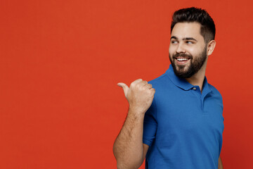 Fototapeta premium Young happy caucasian man 20s wear basic blue t-shirt looking camera point thumb finger aside on workspace area mock up isolated on plain orange background studio portrait. People lifestyle concept.