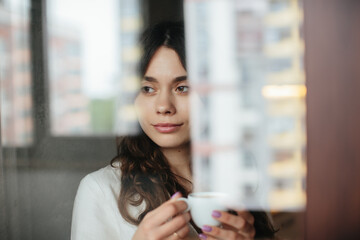 Beautiful caucasian woman drinking hot coffee or tea and looking through window. Indoor background.