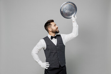 Young fun barista male waiter butler man 20s wearing white shirt vest elegant uniform work at cafe show hold in hand carrying metal tray isolated on plain grey background. Restaurant employee concept.