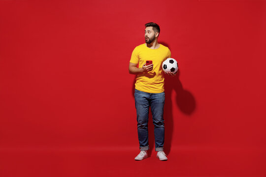 Full Size Body Length Amazed Shocked Surprised Young Bearded Man Football Fan In Yellow T-shirt Look Aside Hold Soccer Ball Use Mobile Cell Phone Isolated On Plain Dark Red Background Studio Portrait.