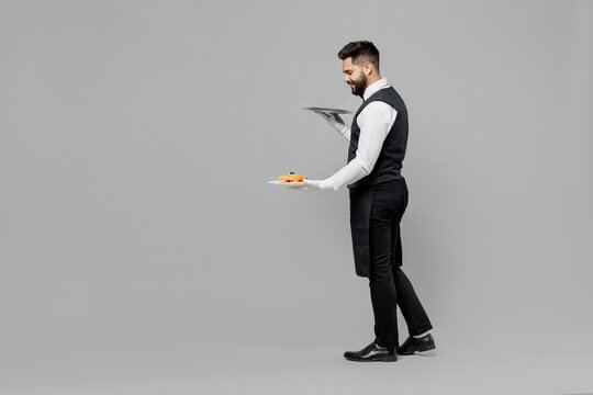 Full size young barista male waiter butler man wear white shirt vest elegant uniform work at cafe hold carry metal tray give cheesecake isolated on plain grey background Restaurant employee concept.