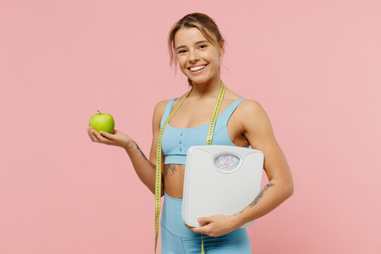 Young sporty athletic fitness trainer instructor woman wear blue tracksuit spend time in home gym hold apple measure tape scales isolated on pastel plain light pink background. Workout sport concept.