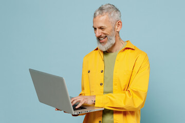 Elderly smiling happy gray-haired mustache bearded man 50s in yellow shirt hold use work on laptop pc computer isolated on plain pastel light blue background studio portrait. People lifestyle concept.