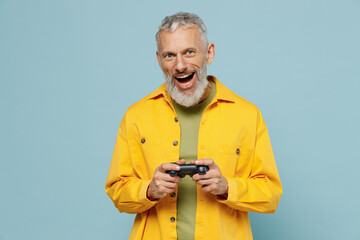 Elderly gray-haired mustache bearded man 50s wear yellow shirt hold in hand play pc game with joystick console isolated on plain pastel light blue background studio portrait. People lifestyle concept.