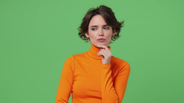 Insighted smart puzzled pensive young brunette woman 20s wears orange shirt looks around thinks scratches at temple comes up with ideas raised finger isolated on plain green background studio portrait