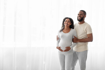 Fototapeta premium Cheerful satisfied handsome young black man hugs pregnant woman with belly in bedroom interior