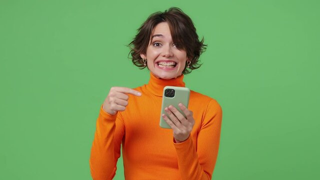 Happy young brunette woman 20s wear orange shirt hold use mobile cell phone typing say wow yes just found out great big win news doing winner gesture isolated on plain green background studio portrait