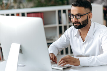 Successful Indian businessman, manager or office worker with beard and glasses, sitting at desk in modern office, wearing white shirt, using computer, texting with colleagues or client, analyzing