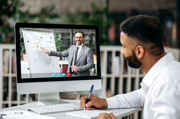 Online business video conference. Concentrated top manager or business owner sitting at desk in office, wearing white shirt, listening online business training, watching coach video, taking notes