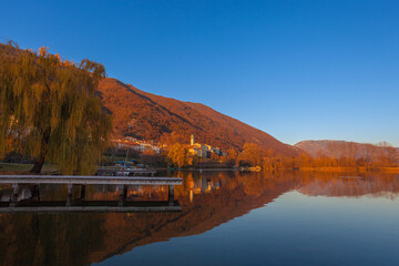 Small village of santa Maria overlooking the lake during an autumn sunset seen with a wooden jetty near a willow tree. Revine Lakes, Veneto, Italy. Concept about reflection and relaxation