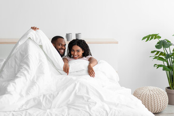 Obraz na płótnie Canvas Glad smiling young black man and woman covered with blanket on bed enjoy happy moment at weekend