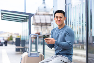 Tourist asian man smiles and looks at the camera sitting at a public transport stop, uses the phone...