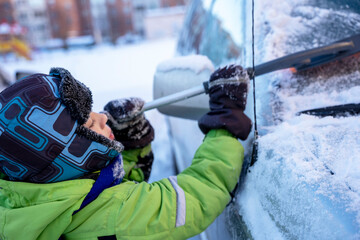 Funny cute Caucasian boy cleans the car from snow with a brush and a scraper