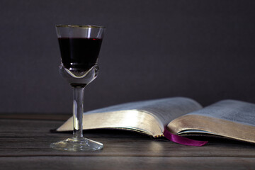 Red wine in a glass and open Holy Bible Book with golden pages on wooden table and grey background.  Copy space for text. A close-up. Christian biblical concept. 