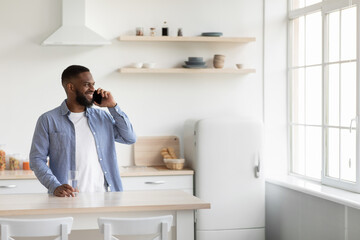 Smiling young african american male with beard and glass of clean water speaks by phone alone