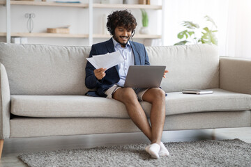 Attractive indian guy sitting on couch, using laptop and headset