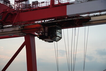 Empty control cabin of red white gantry cranes using for loading and discharging of containers from cargo ships in merchant terminal operated by stevedores. In background blue sky with clouds.