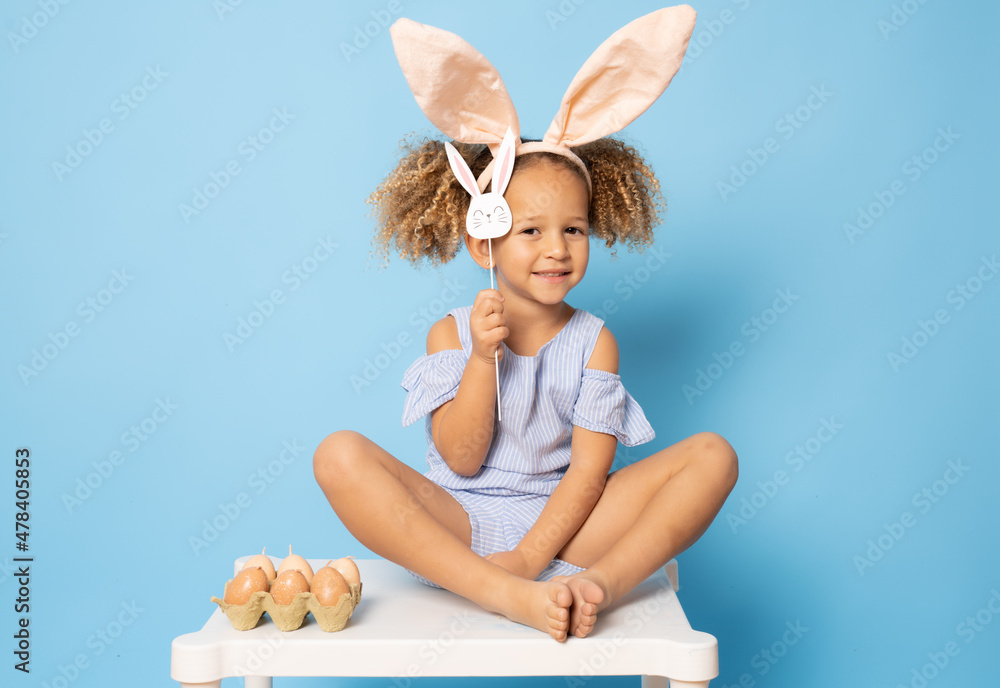 Wall mural cute little girl with curly hair and bunny ears on her head sitting on a table over blue background